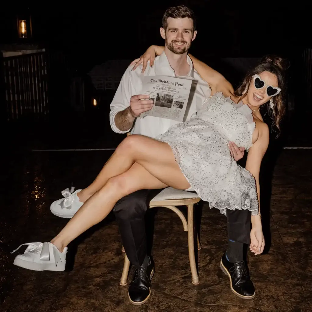 Bride and groom sitting together on a chair holding a just married newspaper outside The Roost Event Center in Mahaffey PA